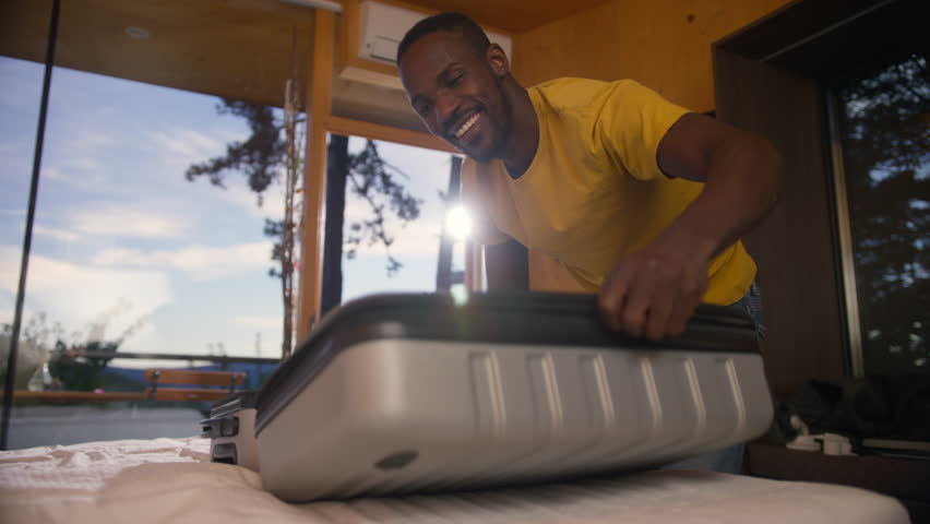 Smiling African American man packing his suitcase in a bedroom, preparing for travel, a long-awaited vacation, handheld shot.