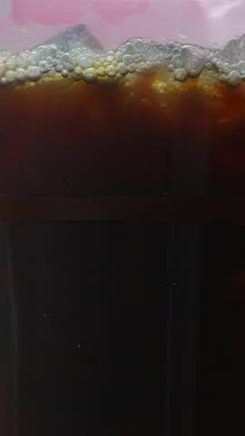 Close-up of iced coffee in a clear cup as milk is poured, creating a dynamic swirl against a soft pink background.