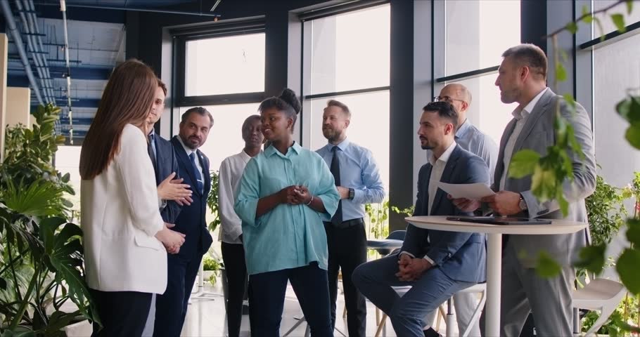 Team of business professionals having a work meeting. Group of multi ethnic people listening to one colleague while standing in a hall with big windows and green plants. Business communication concept