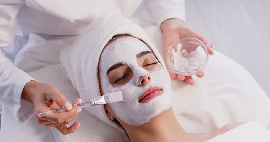 Beautician carefully applies soothing white mask to relaxed womans face with brush at spa. Young Caucasian woman is lying comfortably on massage bed, with her eyes closed. Concept of cosmetology.