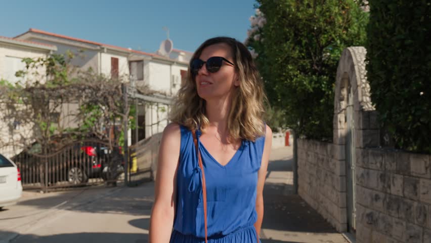 Tourist strolling through the streets of coastal town in Croatia, enjoying sunny weather during summer vacation. Beautiful woman wearing sunglasses and blue dress walks down outdoors