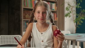 Little Caucasian girl writing copybook eating apple snack break lunch child smile schoolgirl study learning primary education school learner pupil female kid wink bite student healthy food fruit desk - Powered by Shutterstock - Get 15% off with code: PIKWIZARD15