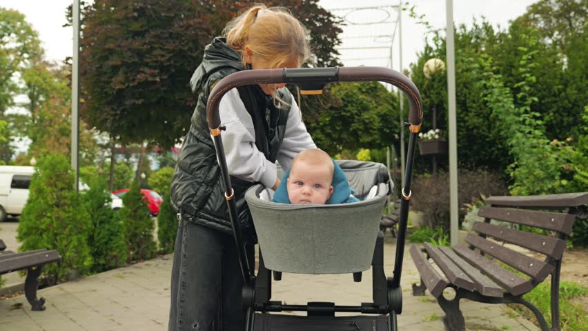 School girl in black down jacket and white jumper playing with a child in sweatshirt who is lying on her stomach in a grey pram. Older sister with baby brother on a walk in the park