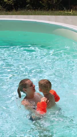 A joyful mother playfully tosses her child wearing orange floaties in a pool, spending family time together on a sunny day. Vertical video 