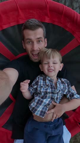 A father and son capture a playful selfie while laughing together on a swing, sharing a joyful and fun outdoor moment.