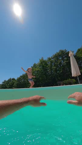 A laughing happy little boy jumps into his daddy's arms and has fun in the swimming pool on a sunny day. Vertical video