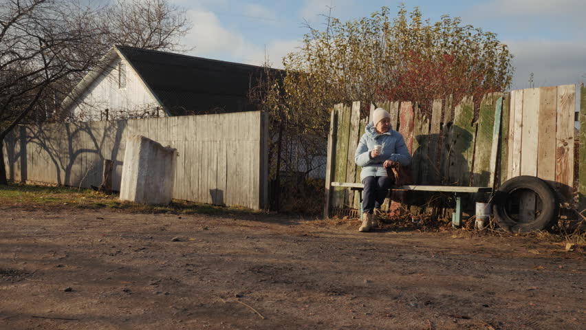 Elderly woman drinking from a cup while sitting on a rustic bench in a peaceful rural setting with a weathered wooden fence behind her. High quality 4k footage