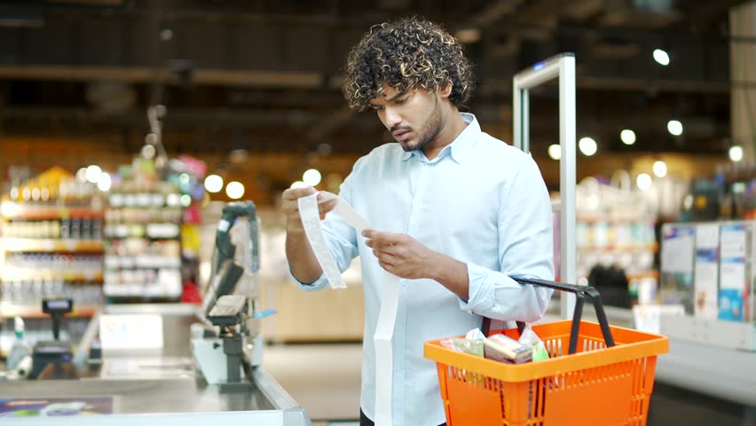 Man customer takes out paper check in grocery store supermarket is surprised at high prices, rising inflation. Gets shocked looking at cashier bill fiscal receipt after shopping dissatisfied shopper