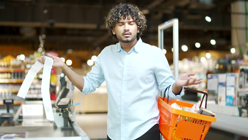 Man customer takes out paper check in grocery store supermarket is surprised at high prices, rising inflation. Gets shocked looking at cashier bill fiscal receipt after shopping dissatisfied shopper