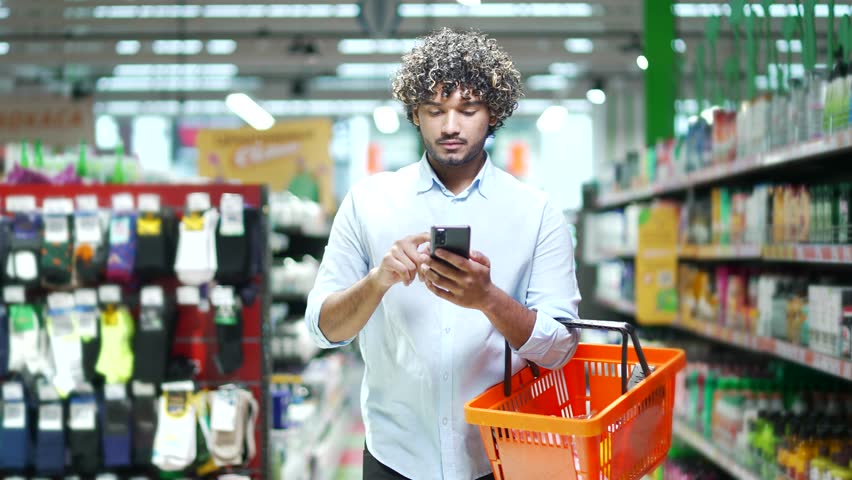 Handsome young man customer checking shopping list using smartphone, purchasing in grocery store in supermarket male buyer shopper pick use mobile phone walk in hypermarket or food market deli