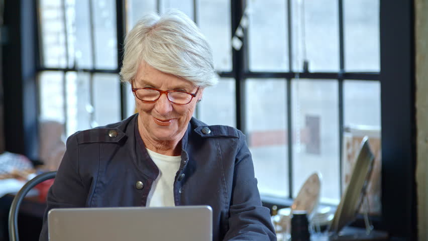 Senior female fashion designer in creative industry sitting at desk working at laptop in studio - shot in slow motion
