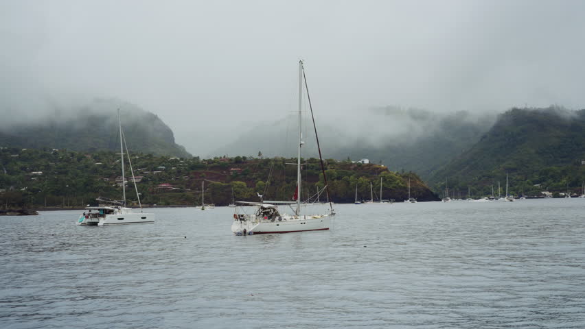 A serene harbor scene unfolds under overcast skies, with mist and clouds enveloping the surrounding hills, as two sailboats are anchored in the calm waters, one prominently in the foreground