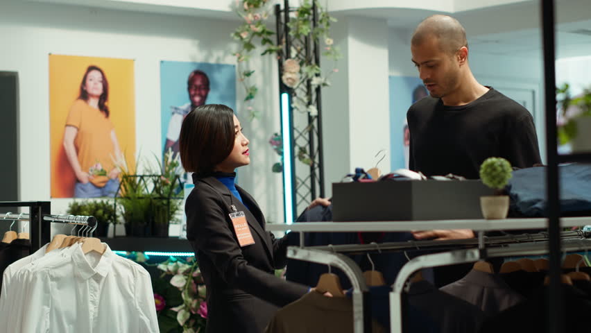 Asian store associate helps a young man client with style advice, embodying expertise and passion and dedication for the fashion industry. Woman improves customer satisfaction. Camera A.