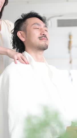 Front view of a Japanese man closing his eyes as his neck and shoulders are massaged at a beauty parlor or salon. Close-up vertical video.