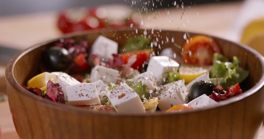A Bowl of Vibrant Mediterranean Greek Salad with Feta Cheese Being Seasoned with Natural Ingredients, Emphasizing Wellness, Balanced Diet, and Fresh Culinary Artistry
