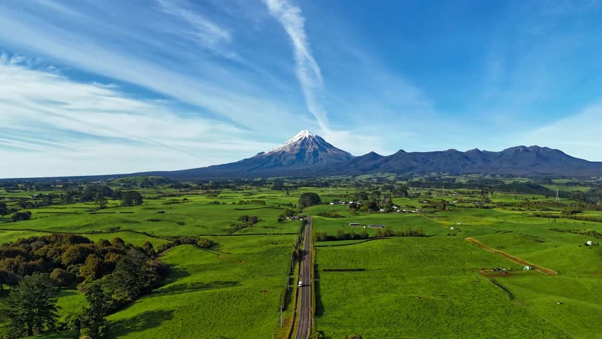 Aerial hyperlapse shot of cars on main street of tropical landscape with Mt. Taranaki in Background. Snowy peak during sunny summer day in New Zealand. Kent Road in NZ. Wide shot.