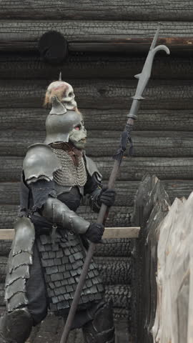 Evil knight stands near burnt wooden house knocking spear on ground. Creature in armor stands at gate to settlement. Monster in medieval village by fence