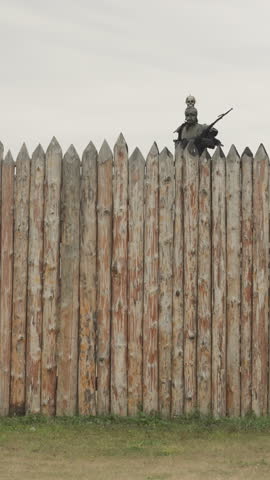 Brave warrior patrols border of settlement walking along high wooden picket fence rain. Medieval sentry in knightly armor inspects territory standing on rise