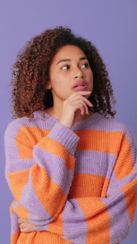 Vertical video, Young woman with curly brown hair wearing striped cozy sweater can't make, spreads arms to sides decision looking at camera on lilac background in studio