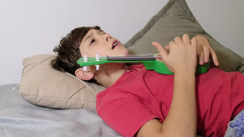 A boy lies on a couch with a green ukulele, his eyes closed as if singing or imagining a melody. This peaceful moment reflects the emotional connection between music and relaxation.