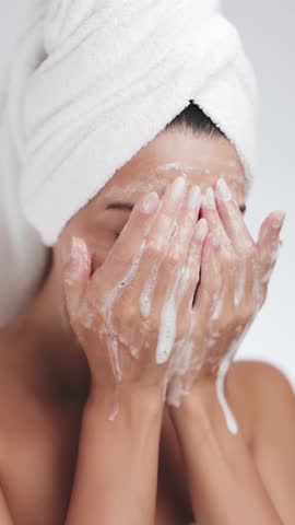 A serene woman wrapped in a towel gently washes her face with foaming cleanser during her skincare routine. Ideal for concepts of beauty, hygiene, self-care, and healthy skin.