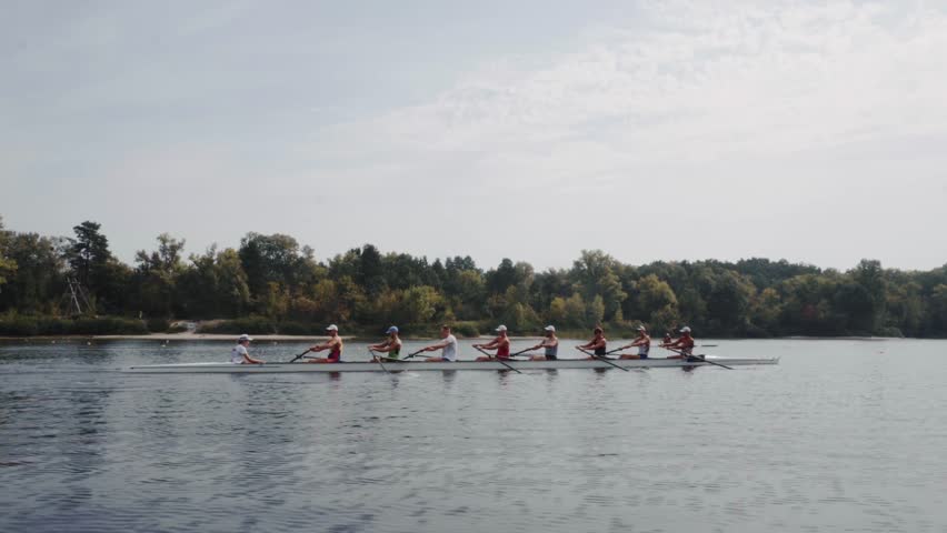 Rowing team training. Side view of 8 young caucasian male rowers, during a rowing practice, athlete sitting in a boat in the river Dnipro, rows through a calm water in autumn. 4k footage. City area in