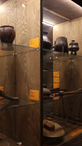 A glass display showcasing traditional pottery, tools, and woven items with detailed labels. The warm wood backdrop enhances the cultural artifacts. This is a vertical video.