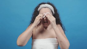 Mature woman removing eye makeup with cotton pads, showing her skincare routine against a vibrant blue backdrop - Powered by Shutterstock - Get 15% off with code: PIKWIZARD15