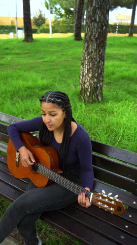 Asian African Young Woman with Brown Skin and Long Black Hair with Dreadlocks Sitting on Bench Playing Guitar in Green Park. Full Shot. Vertical Video
