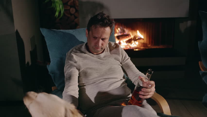 Middle-aged man sitting in a cozy armchair by the fireplace, holding a bottle of alcohol and petting his Golden Retriever. High quality 4k footage