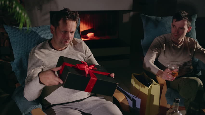 Middle-aged man holding a gift box while sitting in a cozy armchair by a lit fireplace, with his son nearby, celebrating a milestone in a warm home atmosphere. High quality 4k footage