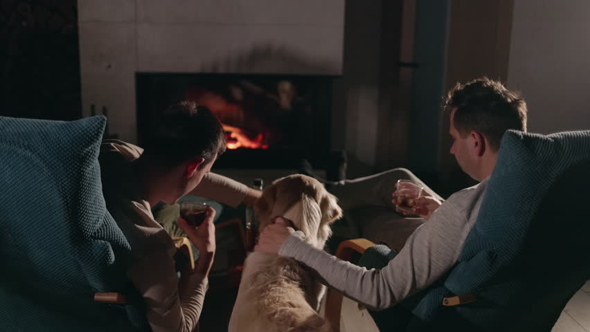 Two men relaxing by the fireplace in the evening, drinking whiskey, with their Golden Retriever nearby, enjoying a cozy and peaceful atmosphere. High quality 4k footage