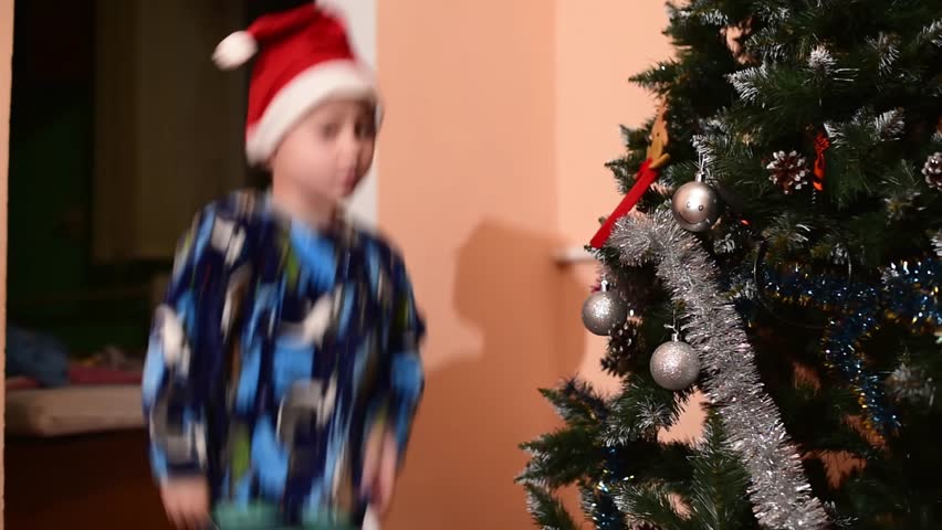 Kid boy in Santa Claus red hat jumps near  Christmas tree, horizontal 4k footage