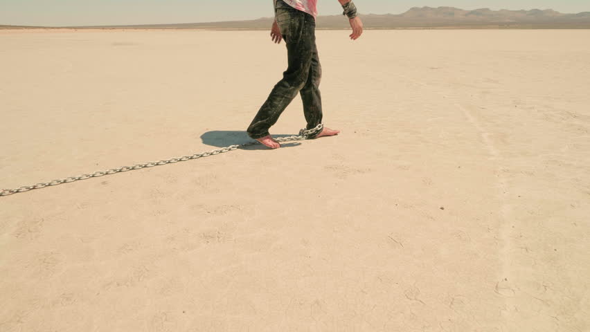 A lone figure walking barefoot across a vast, barren landscape with a heavy chain attached to their ankle. Metaphor of restraint, struggle, inner battle and the will to move on.