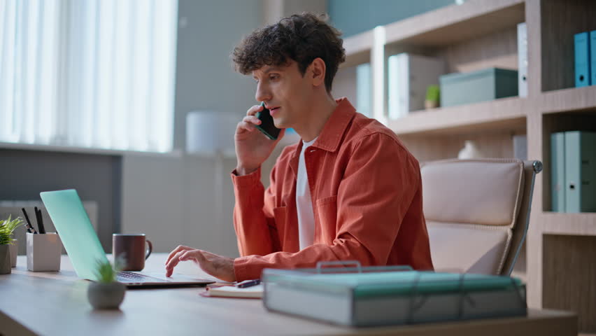 Smiling manager calling in work process sitting office desk. Successful businessman talking smartphone typing laptop at workplace. Happy man entrepreneur making order by cellphone busy of remote job.