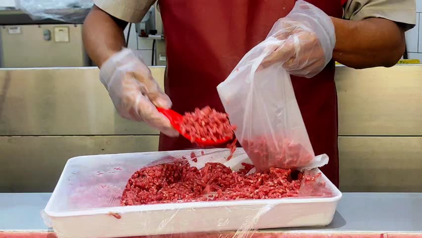 A butcher wearing plastic gloves scooping ground beef into a plastic bag, preparing it for weighing and packaging for a customer in a meat shop.
