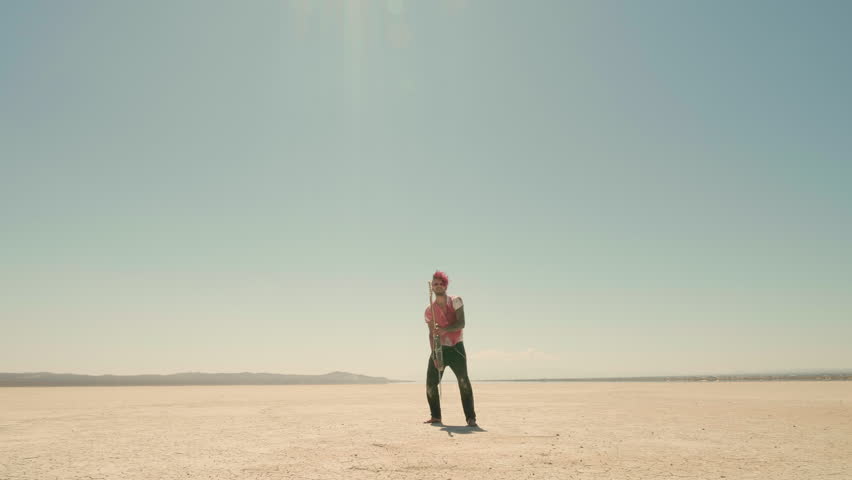 Young man with red dyed hair and blood-covered t-shirt throws an electric guitar up in the air and runs away, letting it freely fall on the scorched earth.
