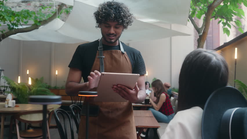 Friendly restaurant worker guy Arabian Indian male waiter in apron serve woman client in cafe customer service smiling man serving in cafeteria communicate with visitor write food order digital tablet