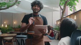 Friendly restaurant worker guy Arabian Indian male waiter in apron serve woman client in cafe customer service smiling man serving in cafeteria communicate with visitor write food order digital tablet - Powered by Shutterstock - Get 15% off with code: PIKWIZARD15