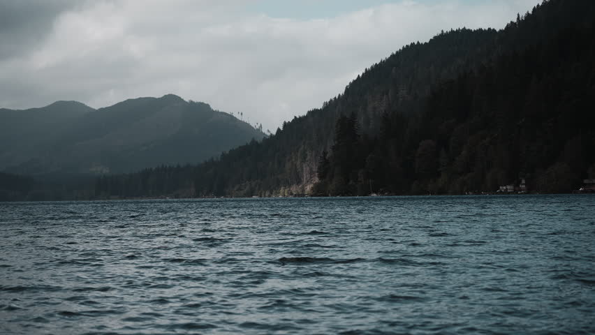 Views of Lake Crescent on a cloudy autumn day. Gentle waves ripple under moody skies, framed by mountains and vibrant fall trees.