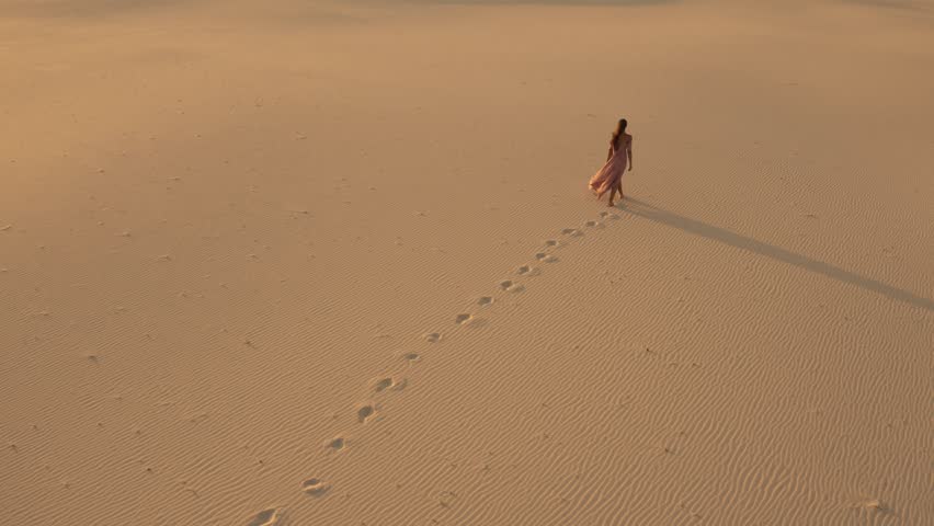 Aerial view from a drone flying next to a gorgeous woman in a pink dress walking on sand dunes in the desert. Traveling fashion girl cinematic footage. 4K Scenic nature