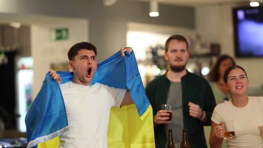 Group of fans of Ukrainian team with a flag are watching the match on TV in a beer bar. They are worried and supporting their favorite team. High quality 4k footage