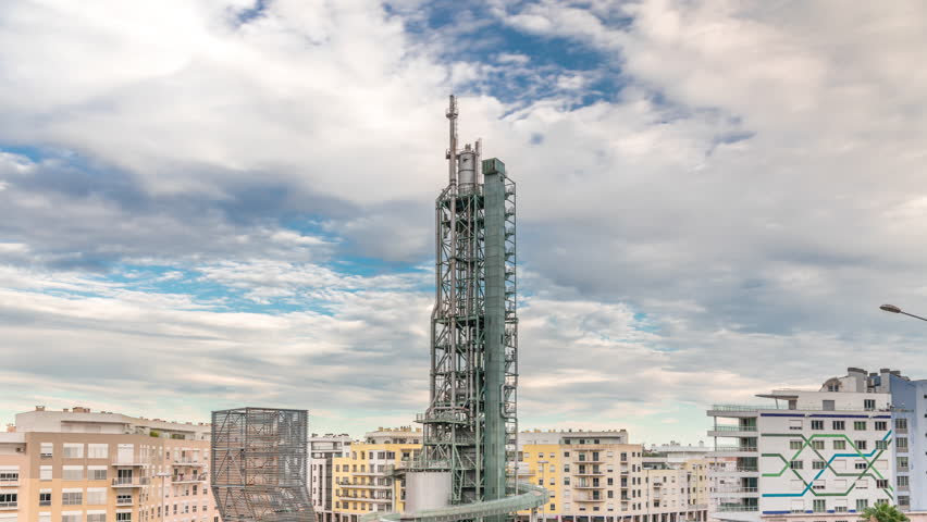 Timelapse hyperlapse of old steel refinery tower with spiral ramp in Lisbon's Park of Nations modern district. Traffic and cloudy sky in the background. Apartment buildings around. Portugal