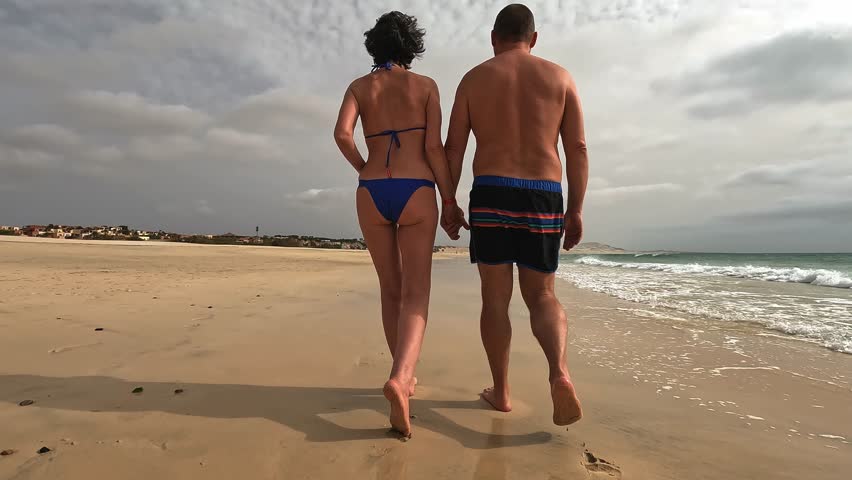 Back view of romantic couple walks hand in hand along peaceful, sandy beach of Boa Vista island with cloudy sky overhead, creating serene and intimate moment by the ocean, Cape Verde