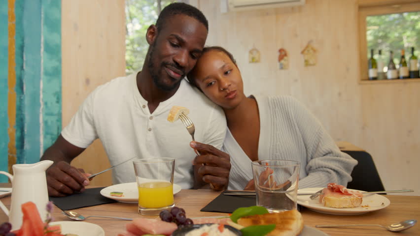 Loving African American couple enjoying daily morning routine on vacation, tasting delicious food for breakfast, in a hotel. Leisure travel concepts.