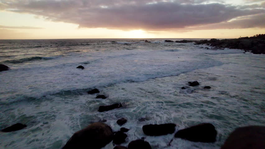 Aerial pull-in of waves crashing along the rocky shoreline in Cape Town, South Africa, moving towards the expansive sea and a vibrant, colorful sunset with clouds on the horizon.