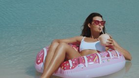 A stylish woman in a white swimsuit relaxes on a colorful inflatable donut with a coconut drink in hand, enjoying the sun by the clear beach waters. Perfect for summer and vacation themes.  - Powered by Shutterstock - Get 15% off with code: PIKWIZARD15