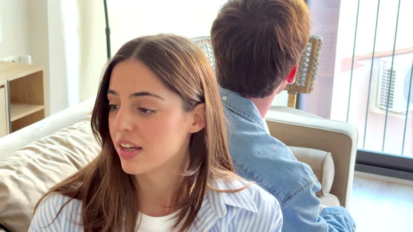 A young couple sitting on a sofa at home, facing emotional distance, with a woman in the foreground and a man turned away, having a heated argument about relationship problems and possible betrayal