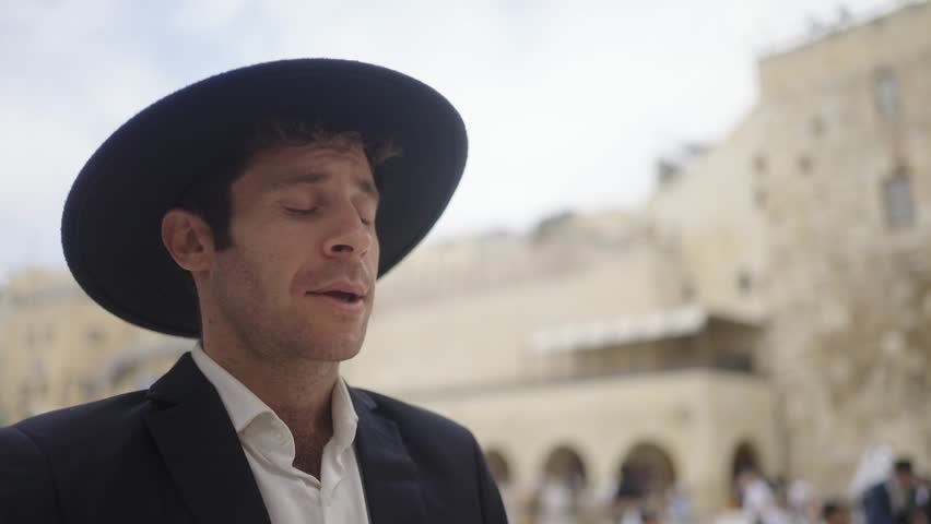 Jewish man praying at the Western Wall in Jerusalem, Israel. He wears a black hat and coat. Stabilized camera moves around subject in slow-motion with selective focus as he makes typical movements.