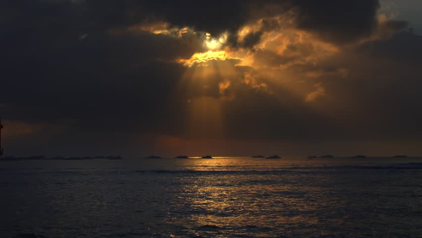 A dramatic sunset over the ocean with rays of sunlight breaking through dark clouds, illuminating the water surface. Silhouettes of boats are visible on the horizon.